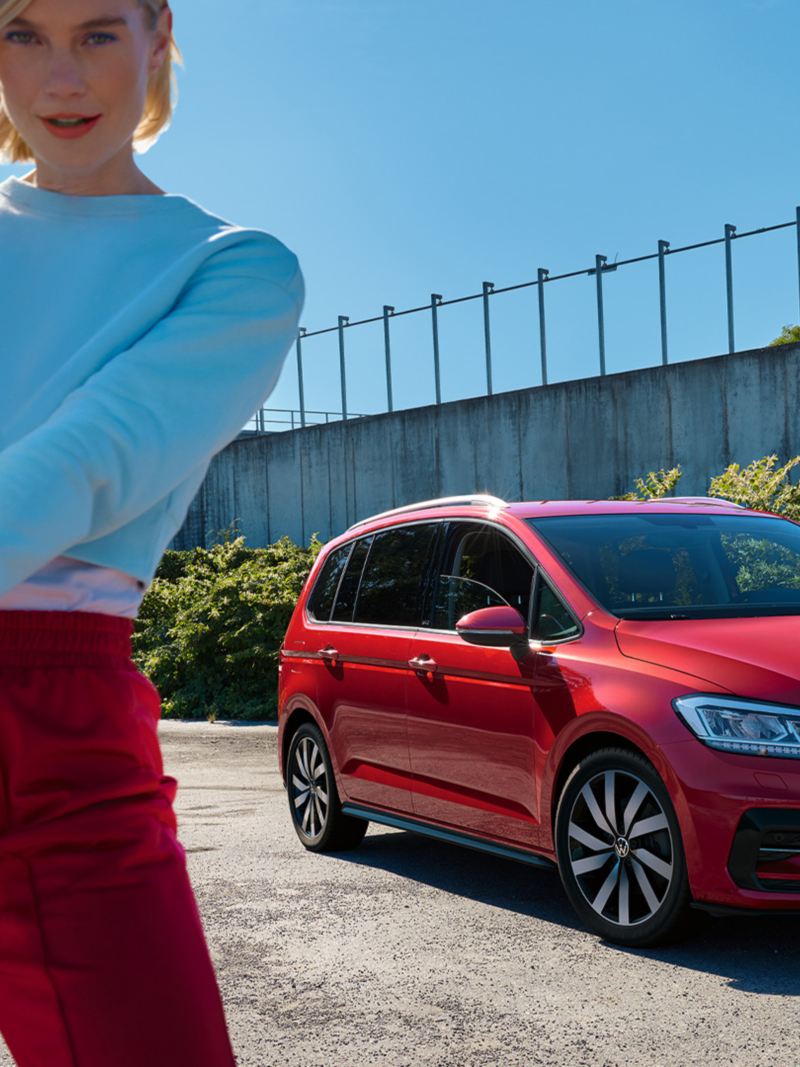 Czerwony VW Touran ACTIVE stoi przed murem z roślinami. W tle niebieskie niebo. Na pierwszym planie kobieta ze srebrną gwiazdą.