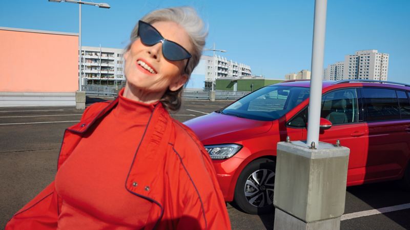 Roter VW Touran ACTIVE auf urbanem Parkplatz. Stadt im Hintergrund. Blick auf Leuchten und Front. Davor Frau mit roter Jacke.