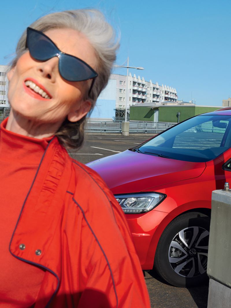 Czerwony VW Touran ACTIVE na parkingu, w tle miasto. Widok na światła i przód nadwozia. Przed nim kobieta w czerwonej kurtce.