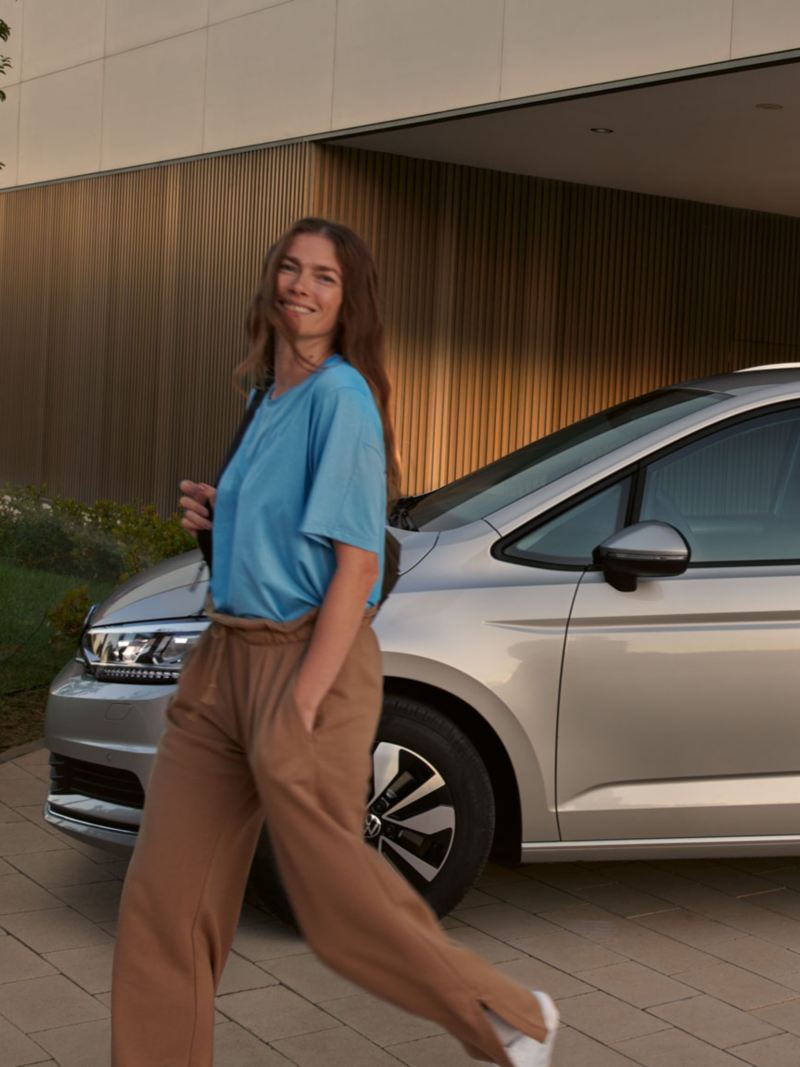 VW Touran MOVE in silber parkt vor einem Gebäude, Ansicht von der Seite außen, eine junge Frau geht lachend daran vorbei