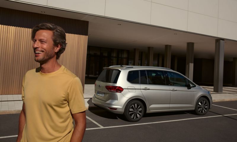 VW Touran MOVE in silber parkt vor einem Gebäude, Blick auf Rückleuchten und Fahrzeugseite, im Vordergrund ein junger Mann 