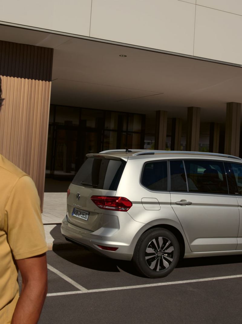 VW Touran MOVE in silber parkt vor einem Gebäude, Blick auf Rückleuchten und Fahrzeugseite, im Vordergrund ein junger Mann 
