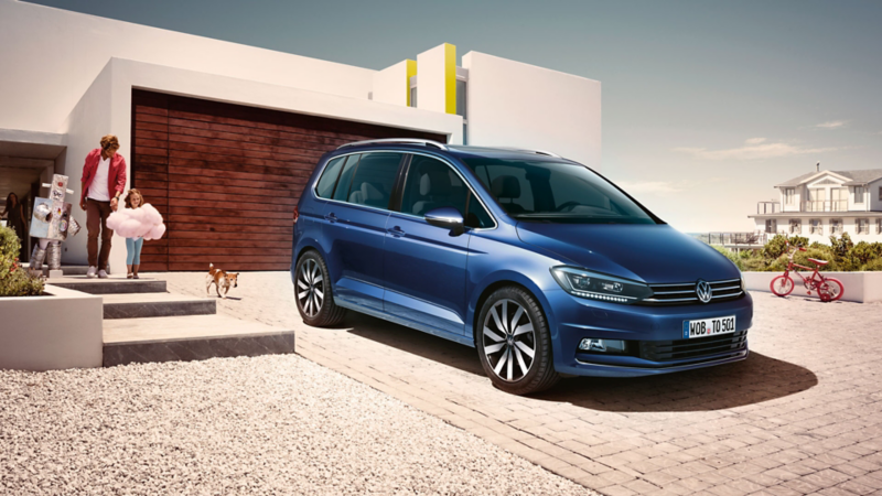 Blauer VW Touran steht auf einer Hausauffahrt, eine Familie mit zwei Kindern und Hund kommt aus dem modernen Haus.