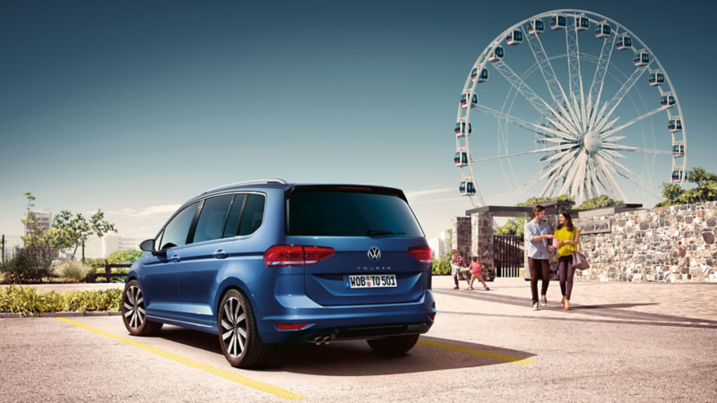 Familie verlässt einen Vergnügungspark mit Riesenrad, im Vordergrund steht ein blauer VW Touran.