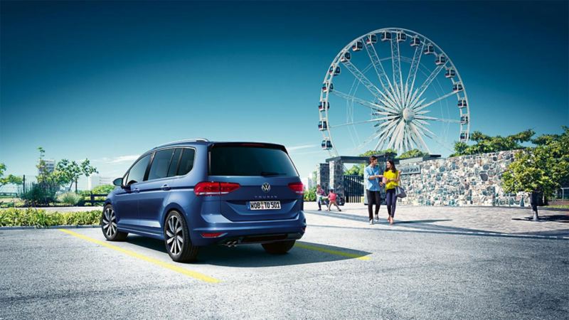 Familie verlässt einen Vergnügungspark mit Riesenrad, im Vordergrund steht ein blauer VW Touran.