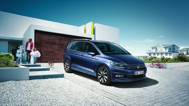 Blue VW Touran in a driveway, a family with two children and a dog come out of the modern house.