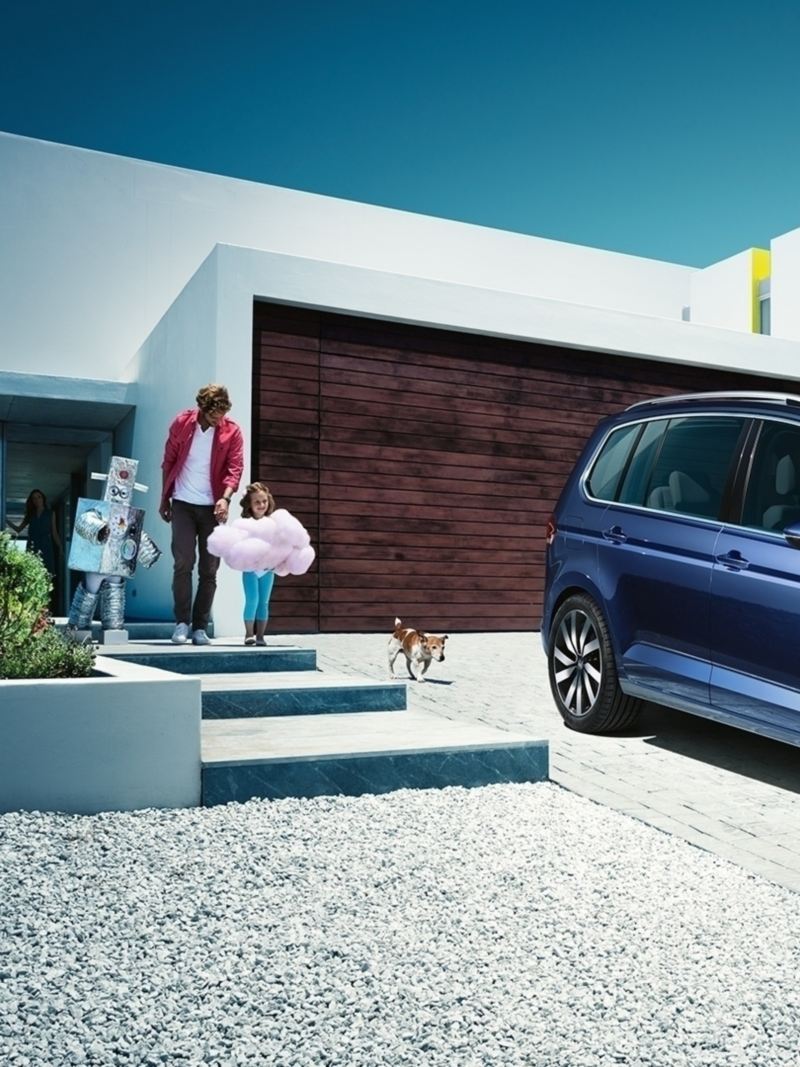 Blauer VW Touran steht auf einer Hausauffahrt, eine Familie mit zwei Kindern und Hund kommt aus dem modernen Haus.