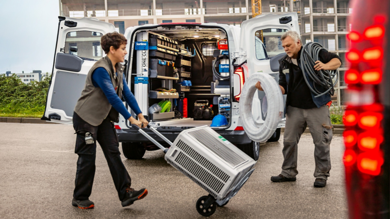 Zwei Personen arbeiten hinter einem Transporter, dessen Hecktüren geöffnet.
