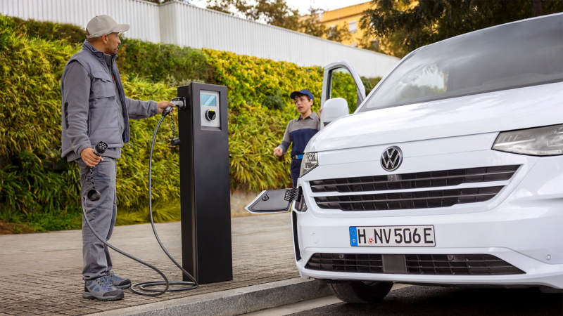 Ein VW e-Transporter wird an einer öffentlichen Ladesäule von einem Handwerker angeschlossen. Aus der Beifahrertür steigt eine zweite Handwerkerin aus.