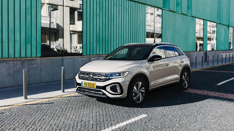 Un Volkswagen T-Roc deux tons, avec une carrosserie beige clair et un toit noir, est garé sur une rue pavée devant un bâtiment moderne à la façade turquoise.
