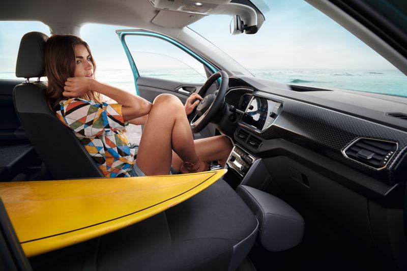 VW T-Cross Cockpit, Frau sitzt auf Fahrersitz bei offener Fahrertür. Auf dem ungeklappten Beifahrersitz liegt ein Surfboard