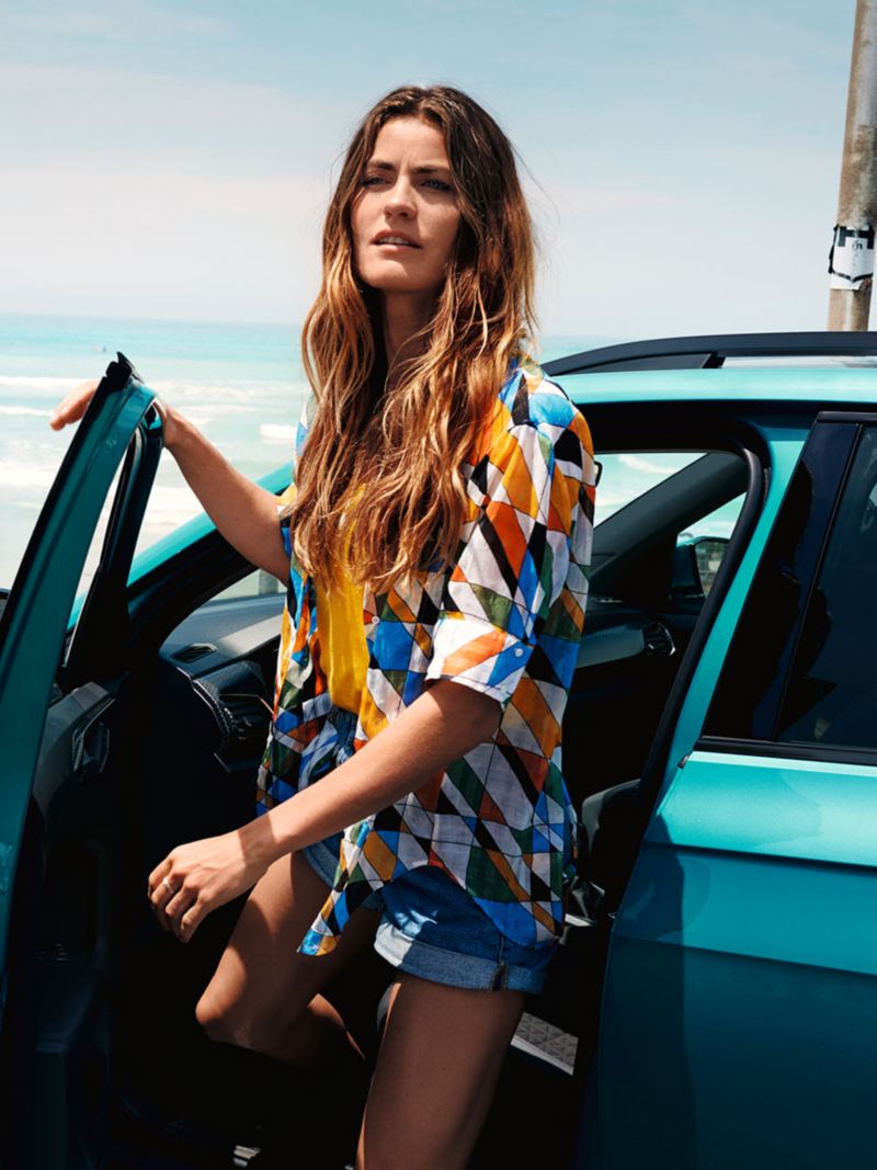 VW T-Cross “R-Line” si trova davanti a una spiaggia e al mare, una donna vestita in abiti estivi esce dal lato conducente.