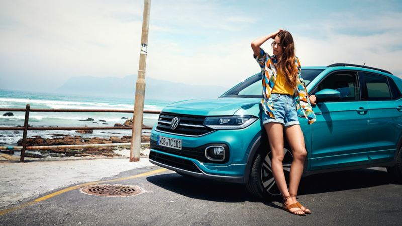 VW T-Cross turquoise stationné devant une plage avec une femme adossé à l'avant du véhicule