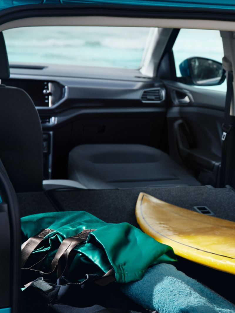 Widok tyłu VW T-Crossa z otwartym bagażnikiem, kobieta wkłada do niego deskę surfingową, złożone prawe przednie siedzenie.