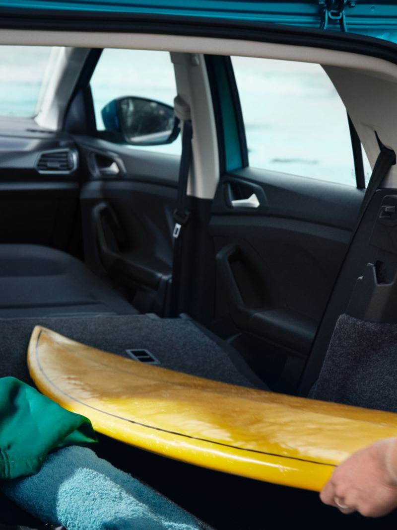 Blick durch geöffnete Heckklappe des VW T-Cross auf den Innenraum, Frau schiebt Surfbrett auf umgeklappten Beifahrersitz.