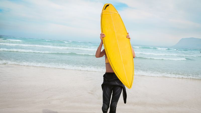 En hvid sandstrand med hav, en kvinde går hen mod den og bærer et surfbræt på ryggen.