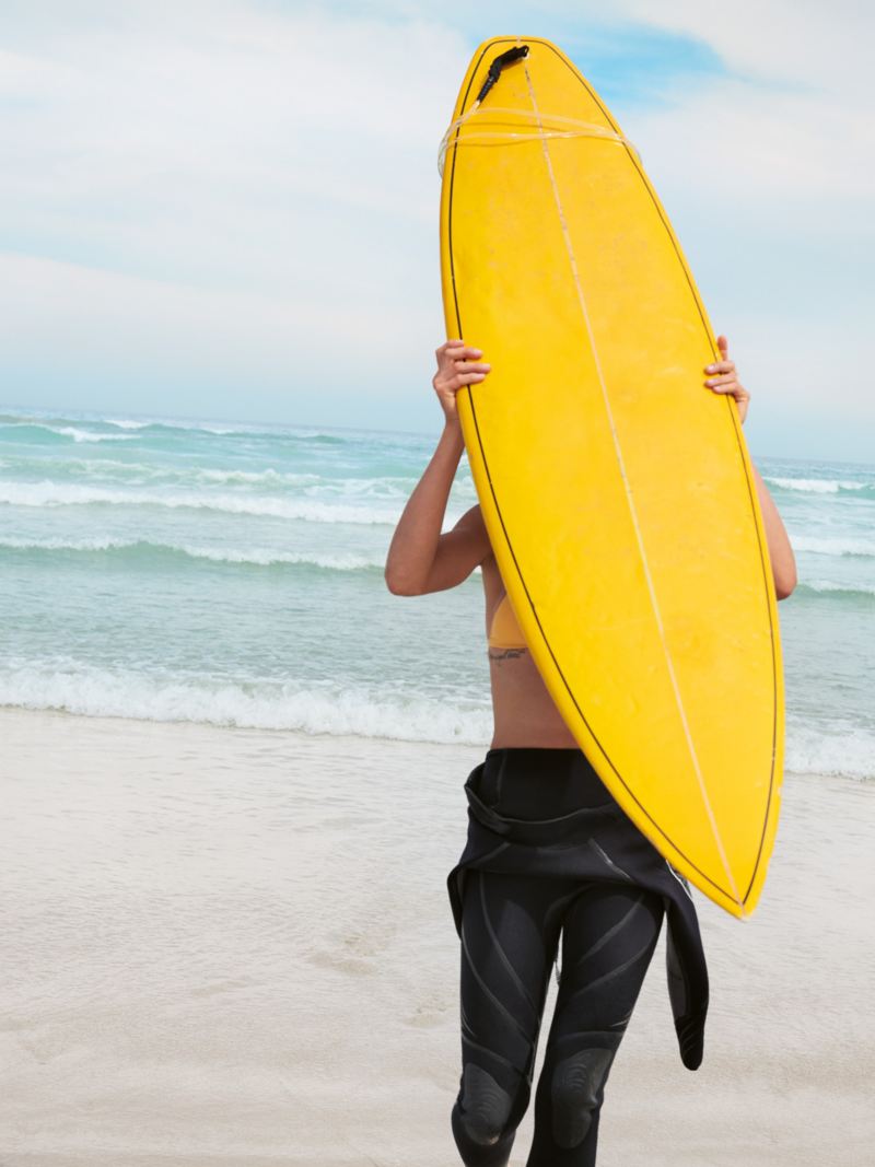 Spiaggia di sabbia bianca, una donna cammina verso il mare portando con sé una tavola da surf sulle spalle.