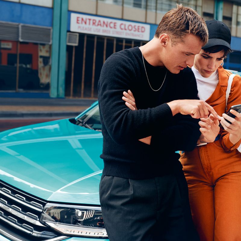 Ein Mann und eine Frau lehnen an der Motorhaube eines parkenden VW T-Cross in Türkis und schauen auf ein Smartphone