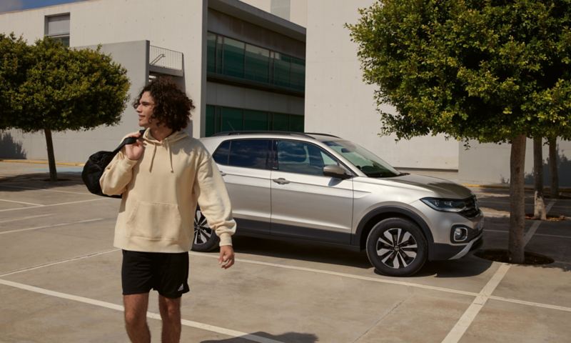 Ein Mann mit einer Sporttasche, im Hintergrund ein parkender VW T-Cross MOVE in silber, Blick auf Frontleuchte und Seite