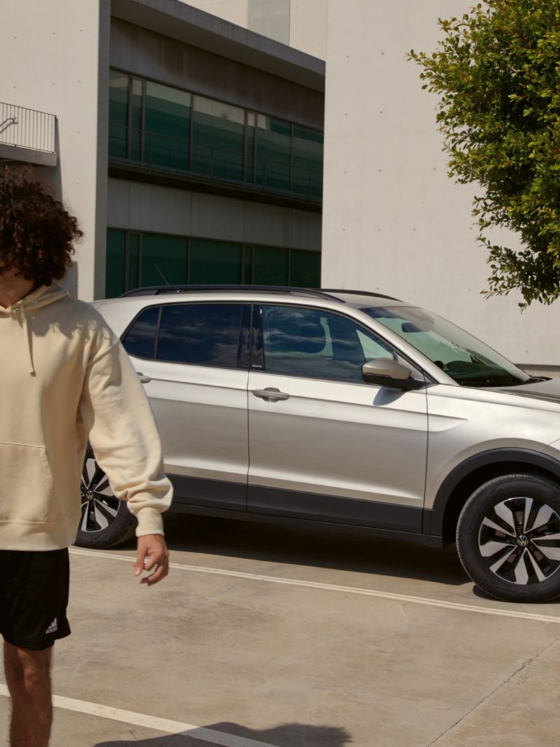 Ein Mann mit einer Sporttasche, im Hintergrund ein parkender VW T-Cross MOVE in silber, Blick auf Frontleuchte und Seite