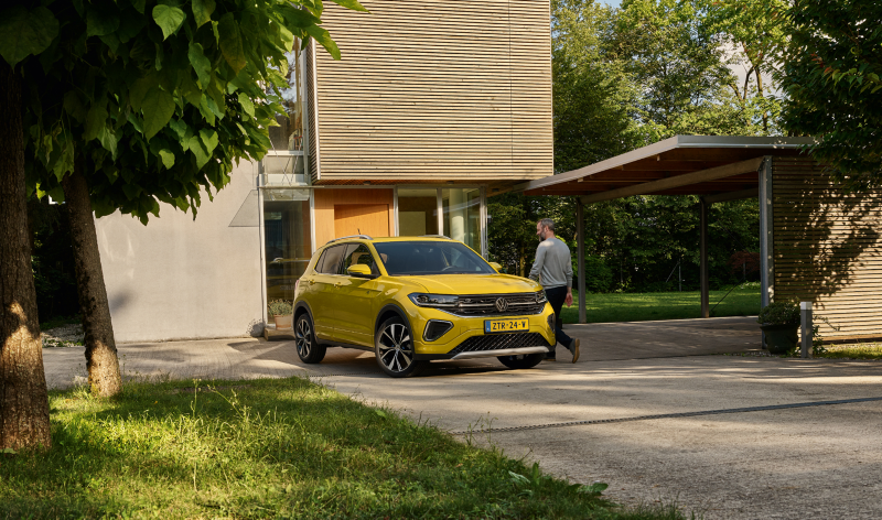 Man loopt naar een gele Volkswagen T-Cross die geparkeerd staat op een oprit, in de omgeving is er veel groen