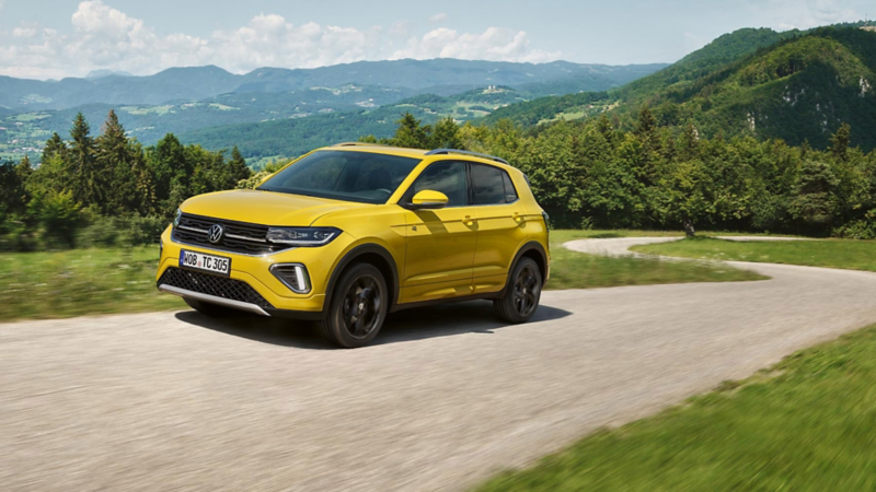En gul VW T-Cross kører på en vej med mange sving gennem et grønt bjerglandskab.