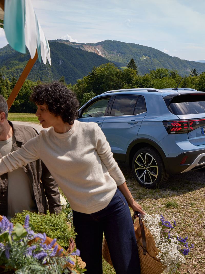 A man and woman stand at a flower stand, behind them a blue VW T-Cross in front of a mountainous landscape.
