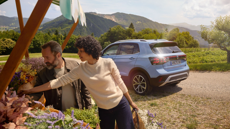 En man och en kvinna står vid ett blomsterstånd, bakom dem en blå VW T-Cross framför ett bergslandskap.