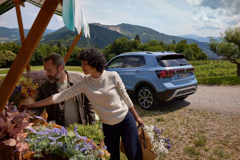 Een man en een vrouw staan bij een bloemenkraampje, achter hen staat een blauwe VW T-Cross voor een bergachtig landschap.