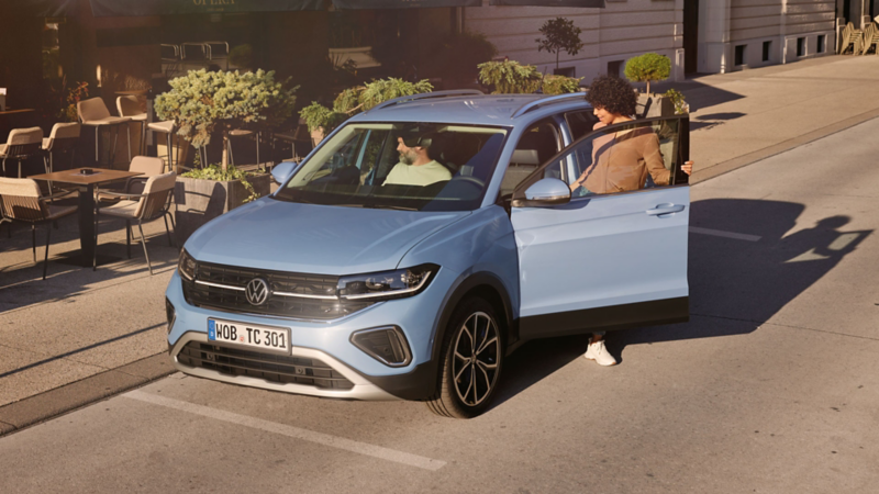 Une femme monte dans un VW T-Cross bleu côté conducteur devant un café.
