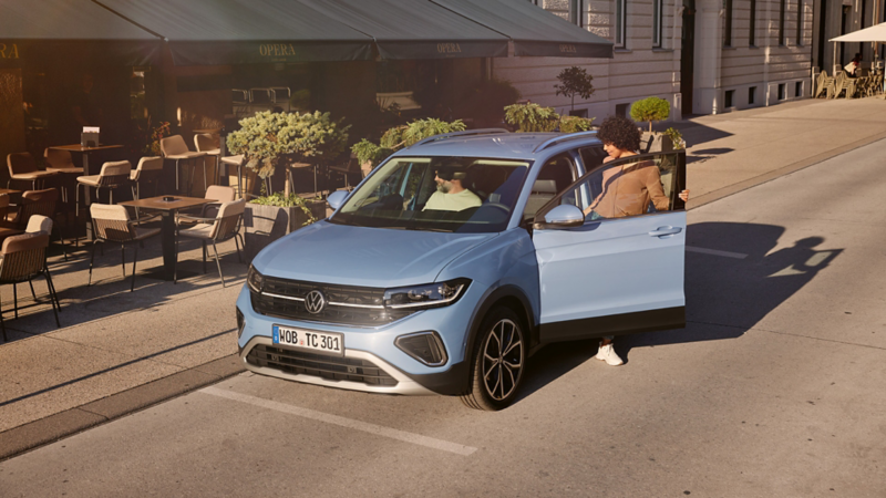 Osoba wysiada z niebieskiego VW T-Cross przed kawiarnią uliczną.
