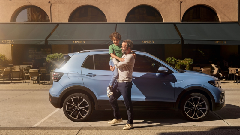 En man håller en glad pojke i sina armar stående framför en blå VW T-Cross.