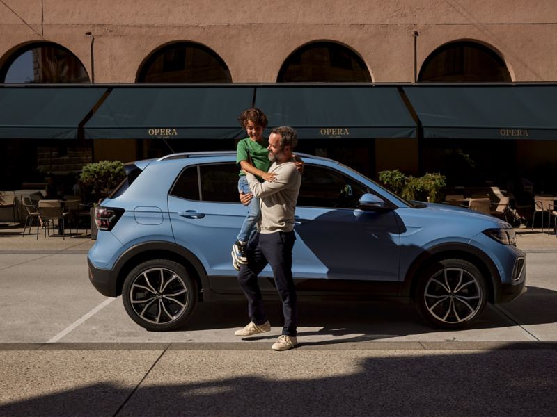 A man holds a happy boy in his arms in front of a blue VW T-Cross.