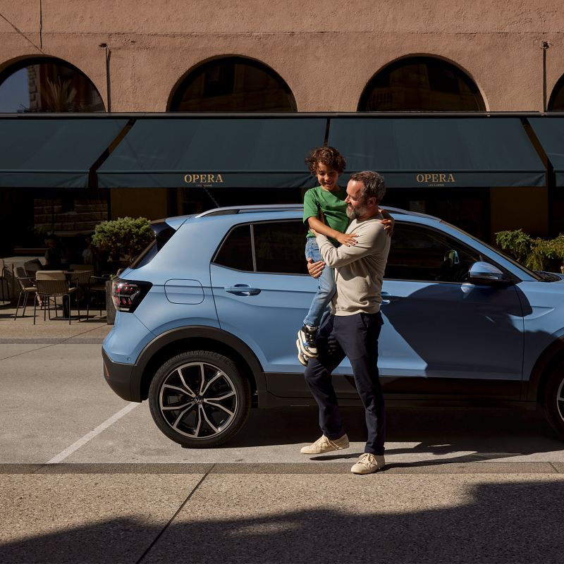 Un homme tient un garçon heureux dans ses bras devant un VW T-Cross bleu.