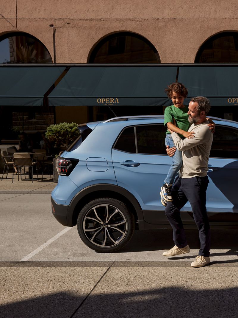 Un homme tient dans ses bras un garçon joyeux devant un VW T-Cross bleu.