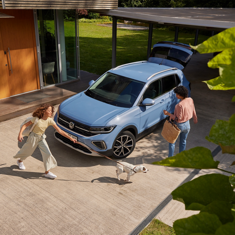 Une fille avec un chien se dirige vers un VW T-Cross bleu dans une allée. La porte passager est ouverte.