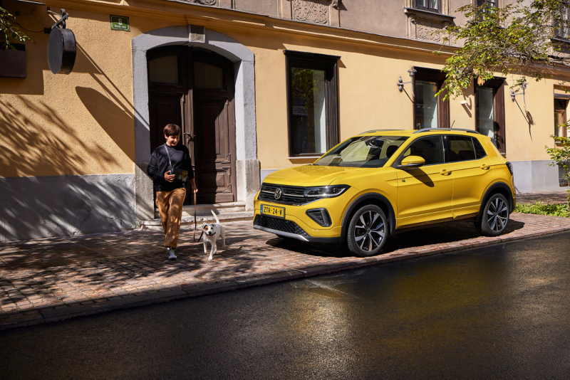 Iemand met hond aan de lijn loopt naast gele Volkswagen T-Cross die geparkeerd staat naast een groot gebouw