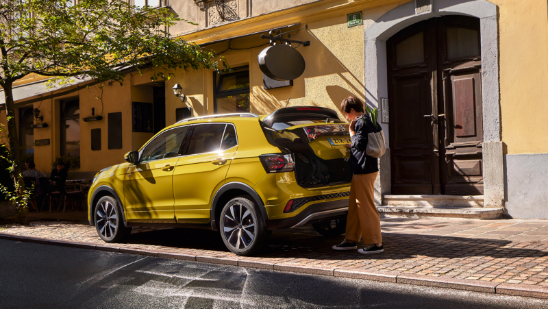 Iemand laad wat in de kofferbak van een gele Volkswagen T-Cross die geparkeerd staat naast een groot gebouw