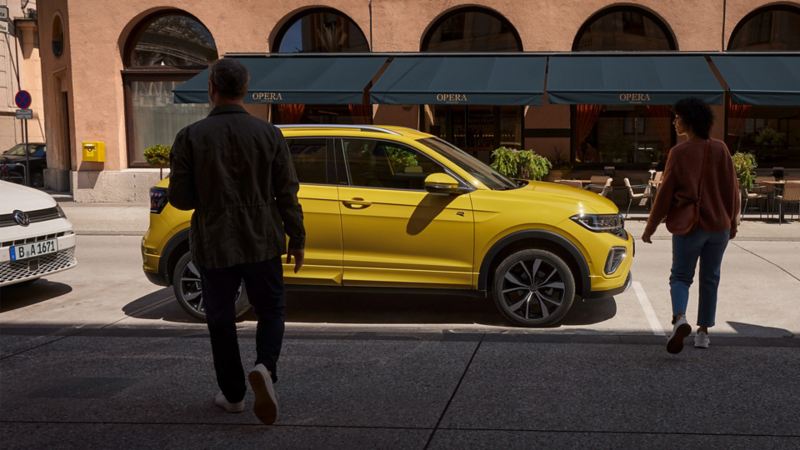 Un homme et une femme s’approchent d’un VW T-Cross jaune sur le bord de la route.