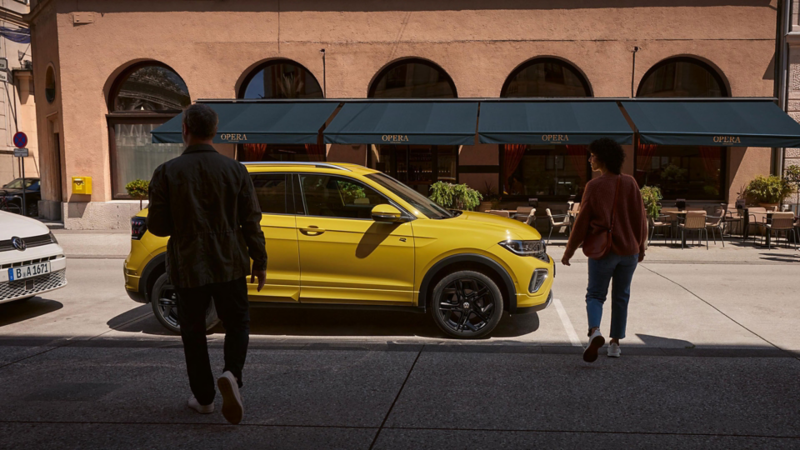 Un homme et une femme s’approchent d’un VW T-Cross jaune sur le bord de la route.