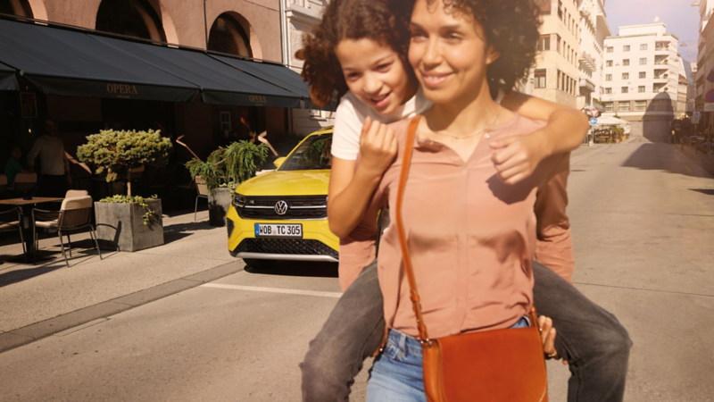 På en gata i stadens centrum bär en kvinna en flicka på ryggen, i bakgrunden syns en gul VW T-Cross.