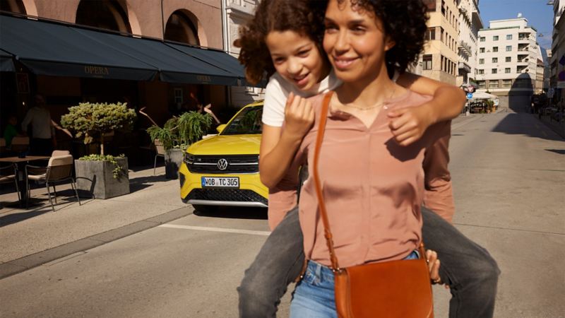 Op een straat in de stadskern draagt een vrouw een meisje op de rug, op de achtergrond staat een gele VW T-Cross.