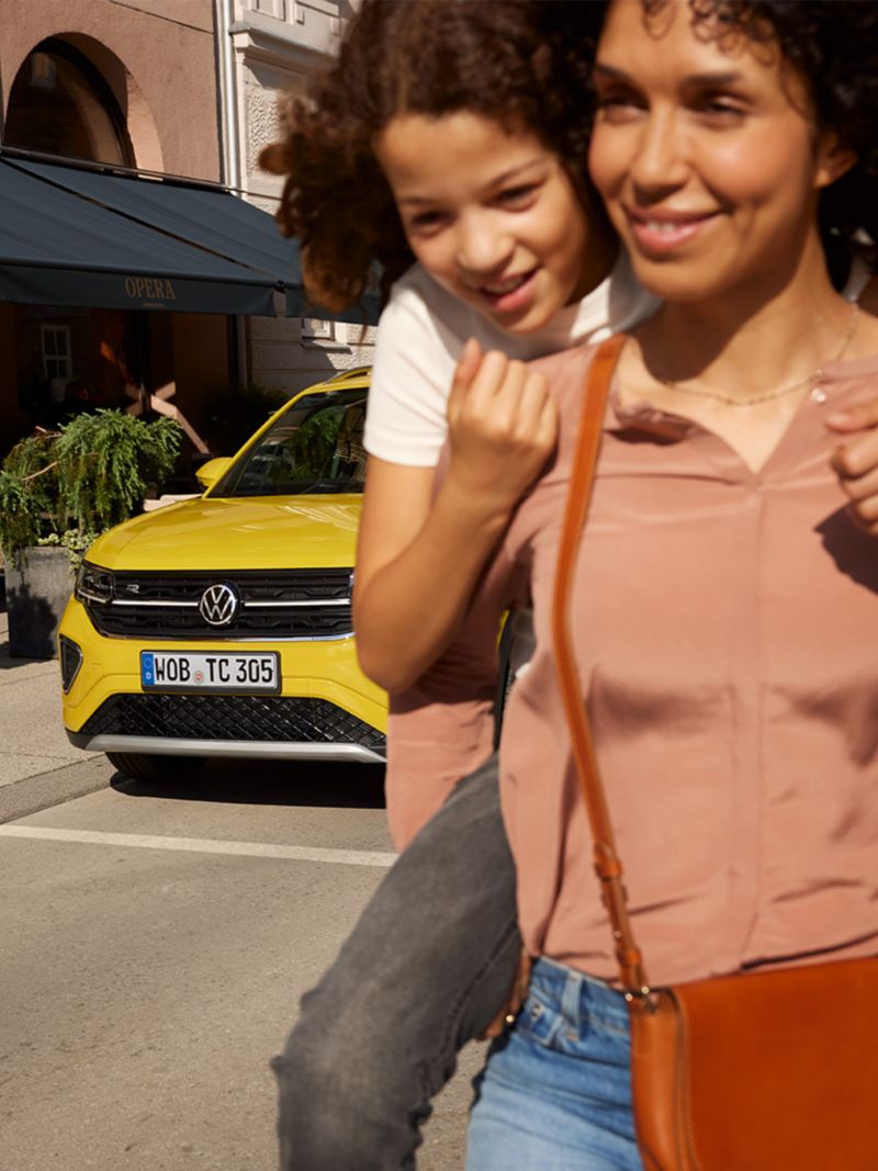 Auf einer Strasse in der Innenstadt trägt eine Frau ein Mädchen auf dem Rücken, im Hintergrund ein gelber VW T-Cross.