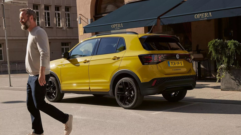 Een man stapt uit een gele Volkswagen T-Cross. Op de achtergrond is het terras van een café te zien.