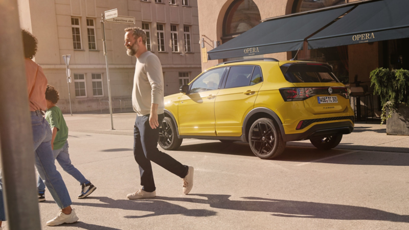Ein gelber VW T-Cross parkt vor einem Café. Eine Familie geht davor über die Straße.