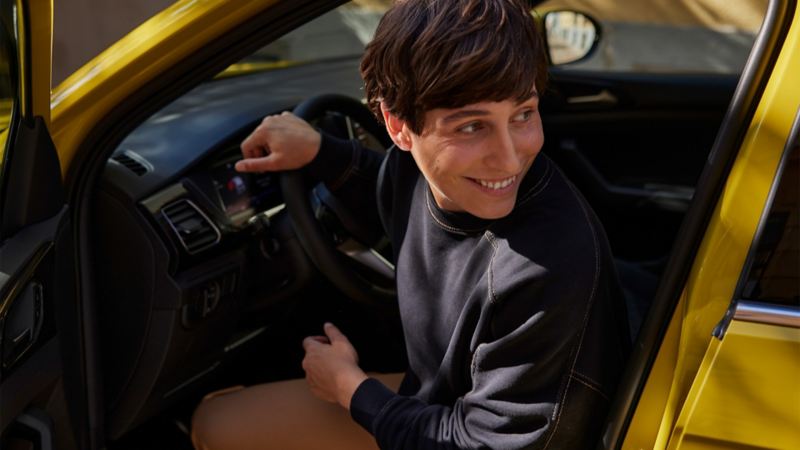Eine junge Frau schaut auf dem Fahrersitz eines gelben VW T-Cross durch die geöffnete Tür lächelnd über die Schulter.