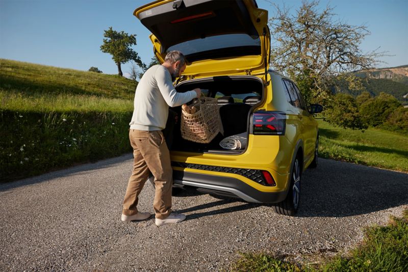 A man puts a large picnic basket in the trunk of a yellow VW T-Cross that is parked on a road in a hilly landscape.