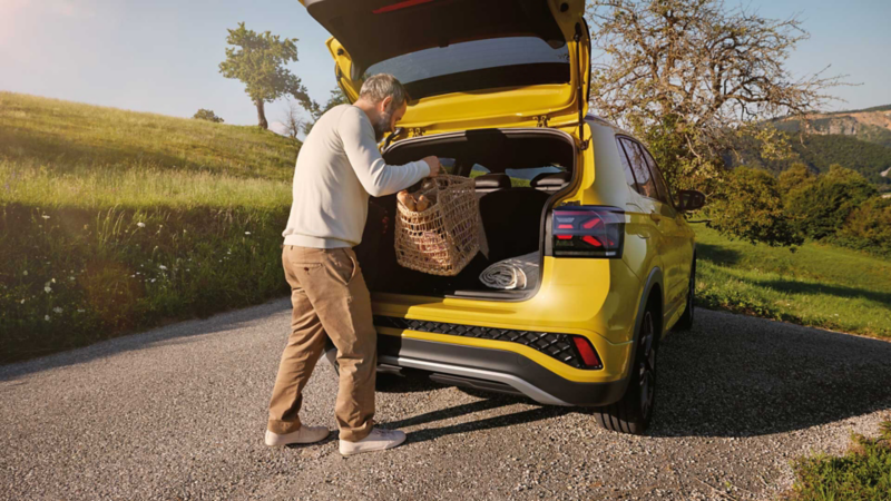 En man lägger en stor picknickkorg i bagageutrymmet på en gul VW T-Cross som står parkerad på en väg i ett kuperat landskap.