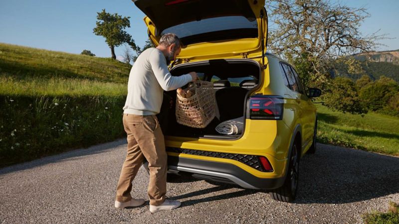 A man puts a large picnic basket in the trunk of a yellow VW T-Cross that is parked on a road in a hilly landscape.