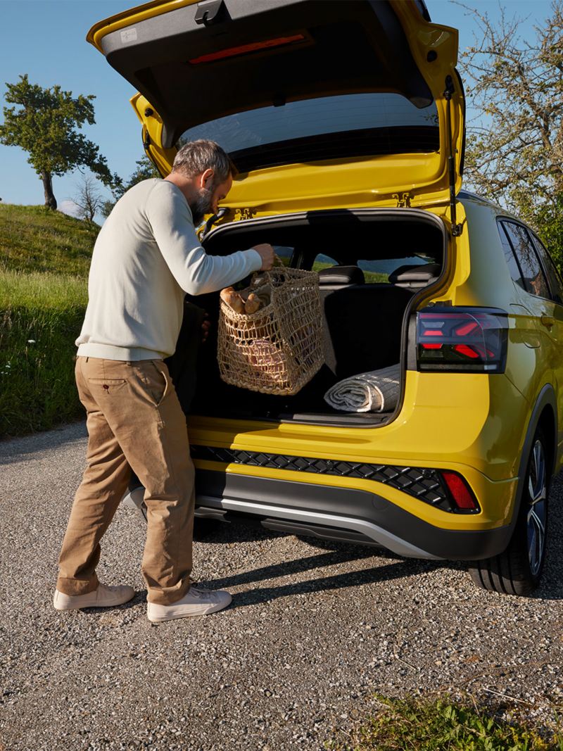 A man puts a large picnic basket in the trunk of a yellow VW T-Cross that is parked on a road in a hilly landscape.